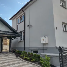 Odnowiona elewacja dwupiętrowego domu z numerem 8, świeżo pomalowana na szaro z białą stolarką okienną, widoczna skrzynka gazowa i oświetlenie zewnętrzne, z zadbanym ogródkiem i brukowanym podjazdem.