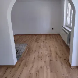 Puste, jasne pomieszczenie z nową, drewnianą podłogą w odcieniach brązu, widoczne białe ściany z łukowym przejściem, grzejnik pod oknem i punktowe oświetlenie sufitowe.