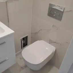 Nowoczesna, podwieszana toaleta z białą ceramiką i chromowanym przyciskiem spłukiwania na ścianie wyłożonej marmurowymi płytkami; obok biała szafka z umywalką i otwór rewizyjny z widocznymi...