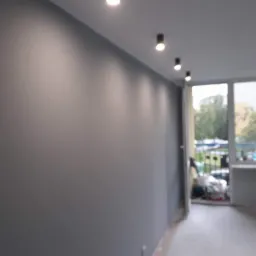 Świeżo pomalowana na szaro ściana z zamontowanymi reflektorami sufitowymi, widok na balkon z zielenią w tle przez duże okno.