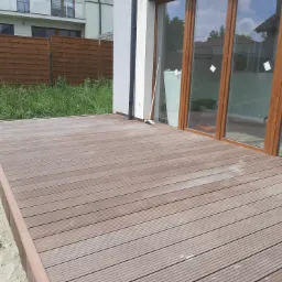 Nowoczesny taras z desek kompozytowych, przylegający do budynku z dużymi oknami, widoczny fragment ogrodu z drewnianym ogrodzeniem.