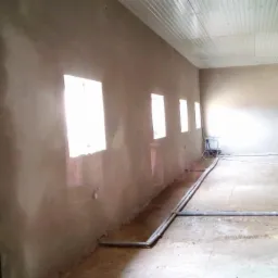 Wnętrze pomieszczenia w trakcie prac wykończeniowych, ściany pokryte świeżym tynkiem, widoczne rury na podłodze i sprzęt do tynkowania w tle.