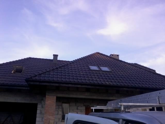 Ciemna dachówka ceramiczna na dachu domu z oknami dachowymi i kominem, częściowo widoczny samochód z drabiną na dachu, błękitne niebo w tle.