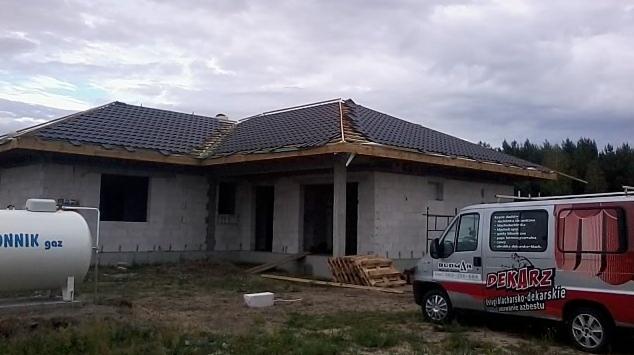 Dom w trakcie budowy, z częściowo położonym, ciemnym dachem z dachówki bitumicznej, widoczny gazowy zbiornik propanu oraz zaparkowany samochód firmy dekarskiej.