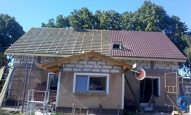 Dom w trakcie prac dekarskich: połowa dachu pokryta łatami, druga połowa czerwoną dachówką z oknem dachowym, widoczne rusztowania i drabiny.