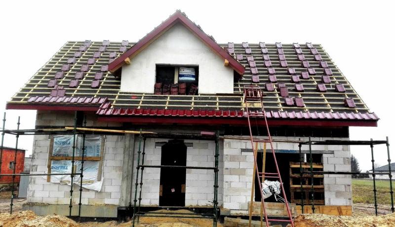 Budowa domu jednorodzinnego w trakcie: widoczne ściany z bloczków, częściowo ułożona dachówka ceramiczna w kolorze ceglastym, rusztowanie i drabina. Dach z oknem dachowym.
