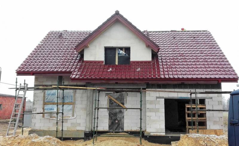 Dom w stanie surowym z nowym, błyszczącym dachem z czerwonej dachówki, widoczne rusztowania i przygotowane materiały budowlane.