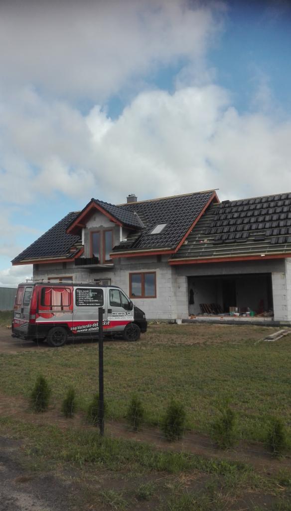 Dom w trakcie budowy z częściowo pokrytym czarną dachówką dachem, na podwórku stoi srebrny bus firmy dekarskiej z reklamą na boku.