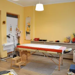 Jasne wnętrze gabinetu z żółtymi ścianami, wyposażone w stół do masażu z czerwonym obramowaniem i białym prześcieradłem, drewniane schodki, lampę na statywie, komodę z dekoracjami i wiszący obraz...