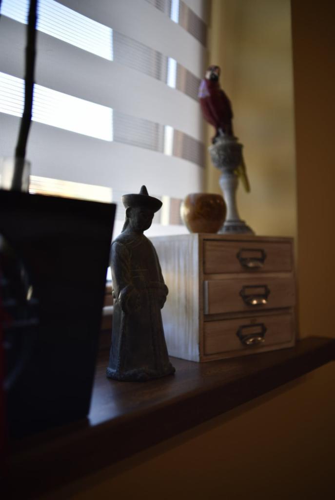 Dekoracyjny kącik z figurką w chińskim stylu, drewnianą komódką z szufladami i figurką papugi na tle żaluzji.