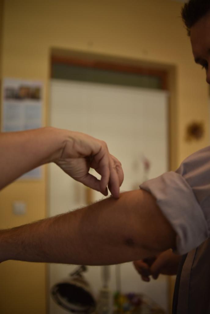 Dłoń terapeuty dotyka ramienia pacjenta w trakcie sesji terapeutycznej, widoczny cień na ścianie i fragment ubioru pacjenta.