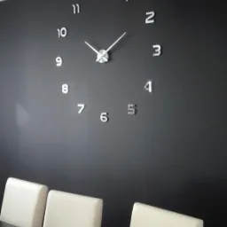 Ściana w ciemnym kolorze z dużym, srebrnym zegarem naklejanym, trzy białe krzesła przy ciemnym stole, widoczny fragment minimalistycznego wnętrza.