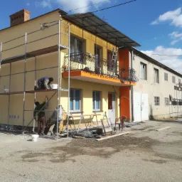 Dwóch pracowników na rusztowaniu maluje elewację dwupiętrowego budynku na kolor żółty, widoczny balkon z ozdobną, kutą balustradą i pomarańczowym podbiciem, na ziemi leżą materiały budowlane.