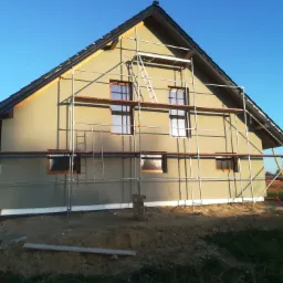 Dom jednorodzinny w trakcie prac wykończeniowych elewacji, widoczne rusztowania i nałożona warstwa tynku zewnętrznego. Okna częściowo zasłonięte. W tle błękitne niebo.