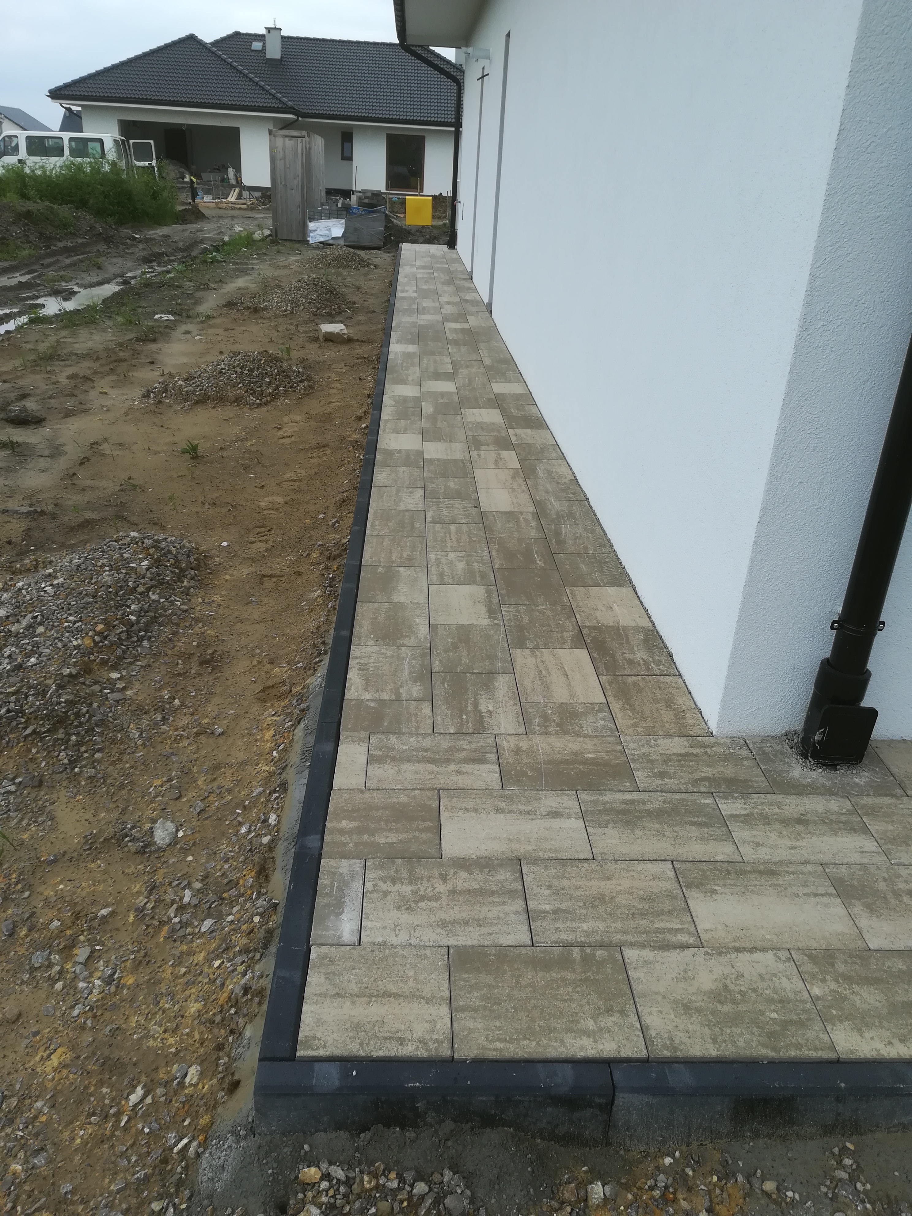 Świeżo ułożona ścieżka z kostki brukowej przy białej ścianie domu, widoczne czarne obrzeża i teren przygotowany pod dalsze prace.