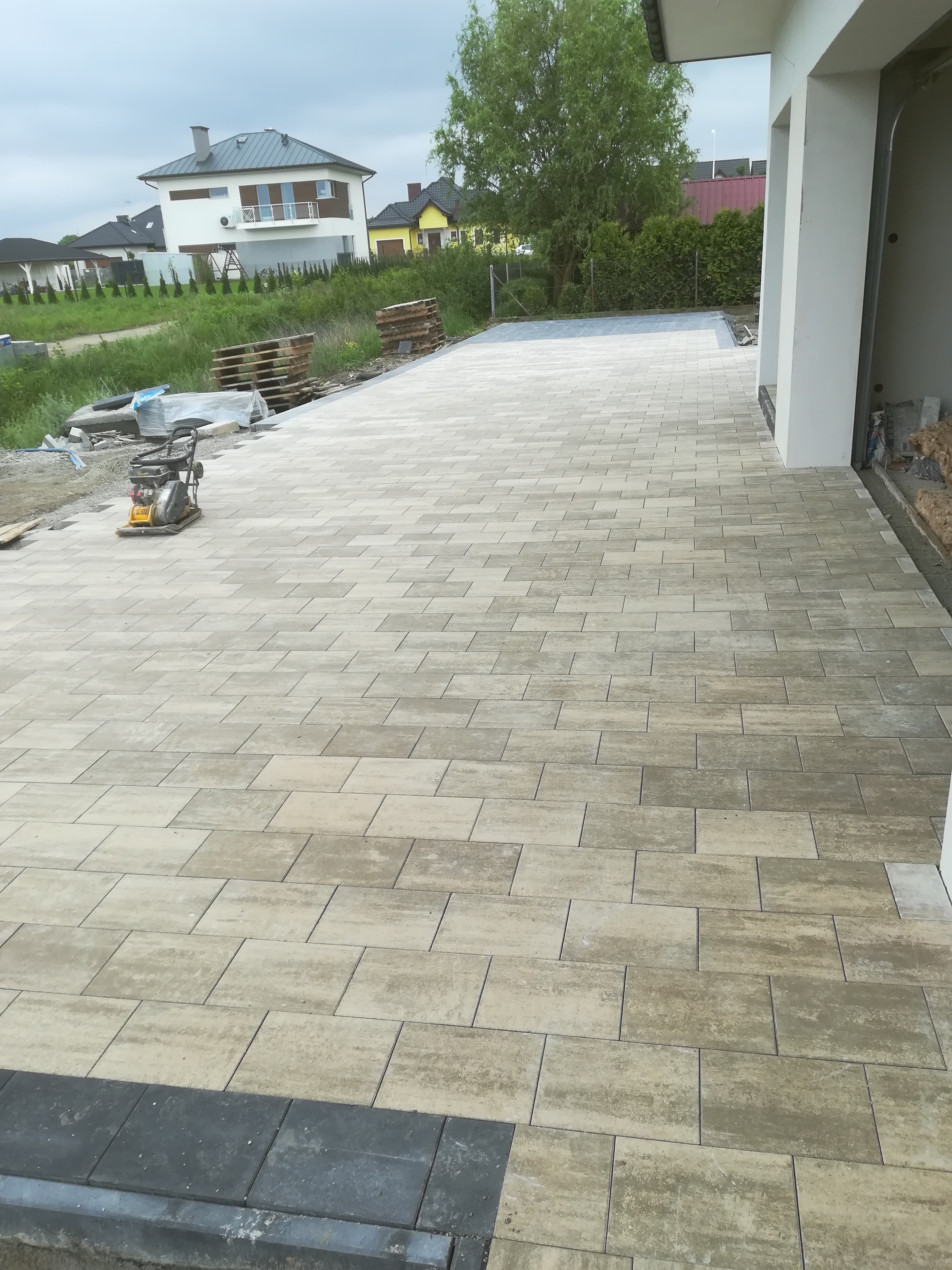 Ułożona kostka brukowa na podjeździe z widoczną ubijarką w tle, paletami kostki i domami jednorodzinnymi.