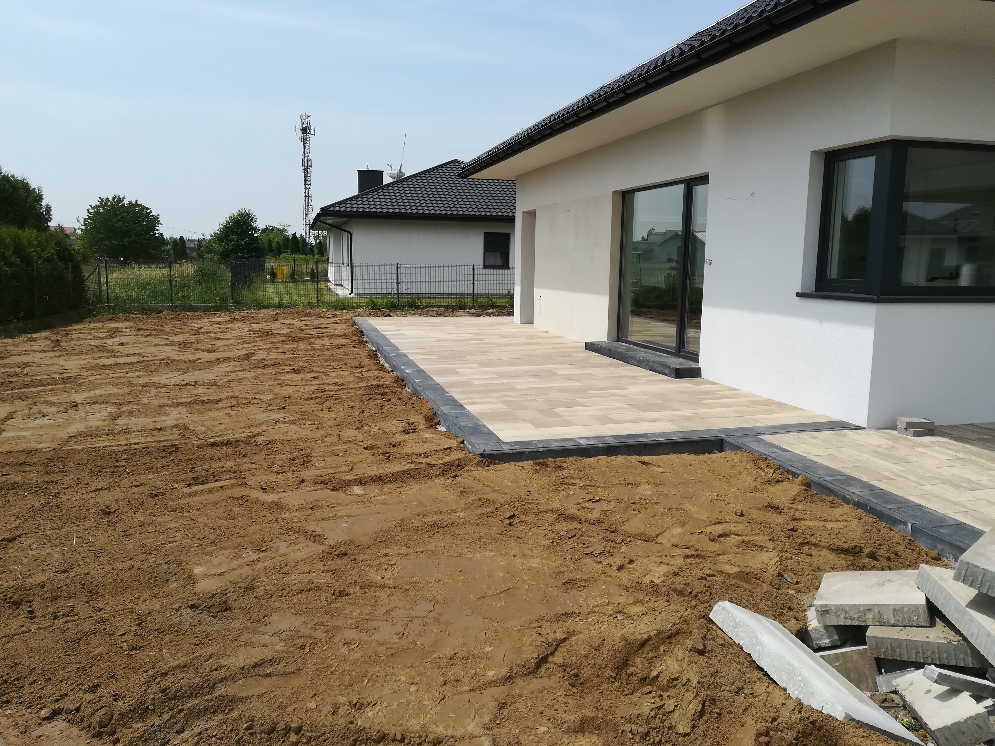 Jasna kostka brukowa z ciemną obwódką na tarasie przylegającym do białego domu, widok na przygotowany teren pod dalsze prace brukarskie, stos kostki w rogu.