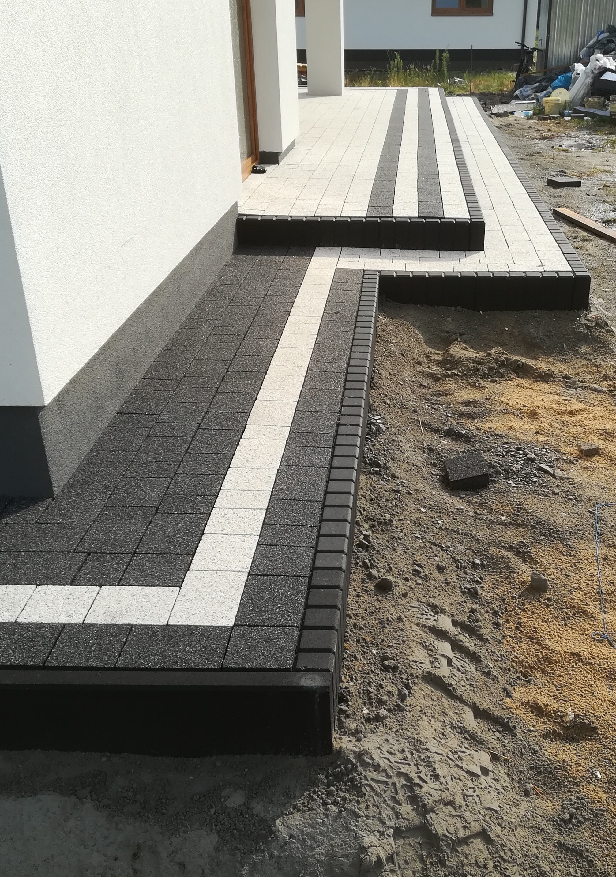 Ułożona kostka brukowa przed domem, z jasnym pasem wzdłuż ścieżki i schodami, widoczne ślady w piasku obok.