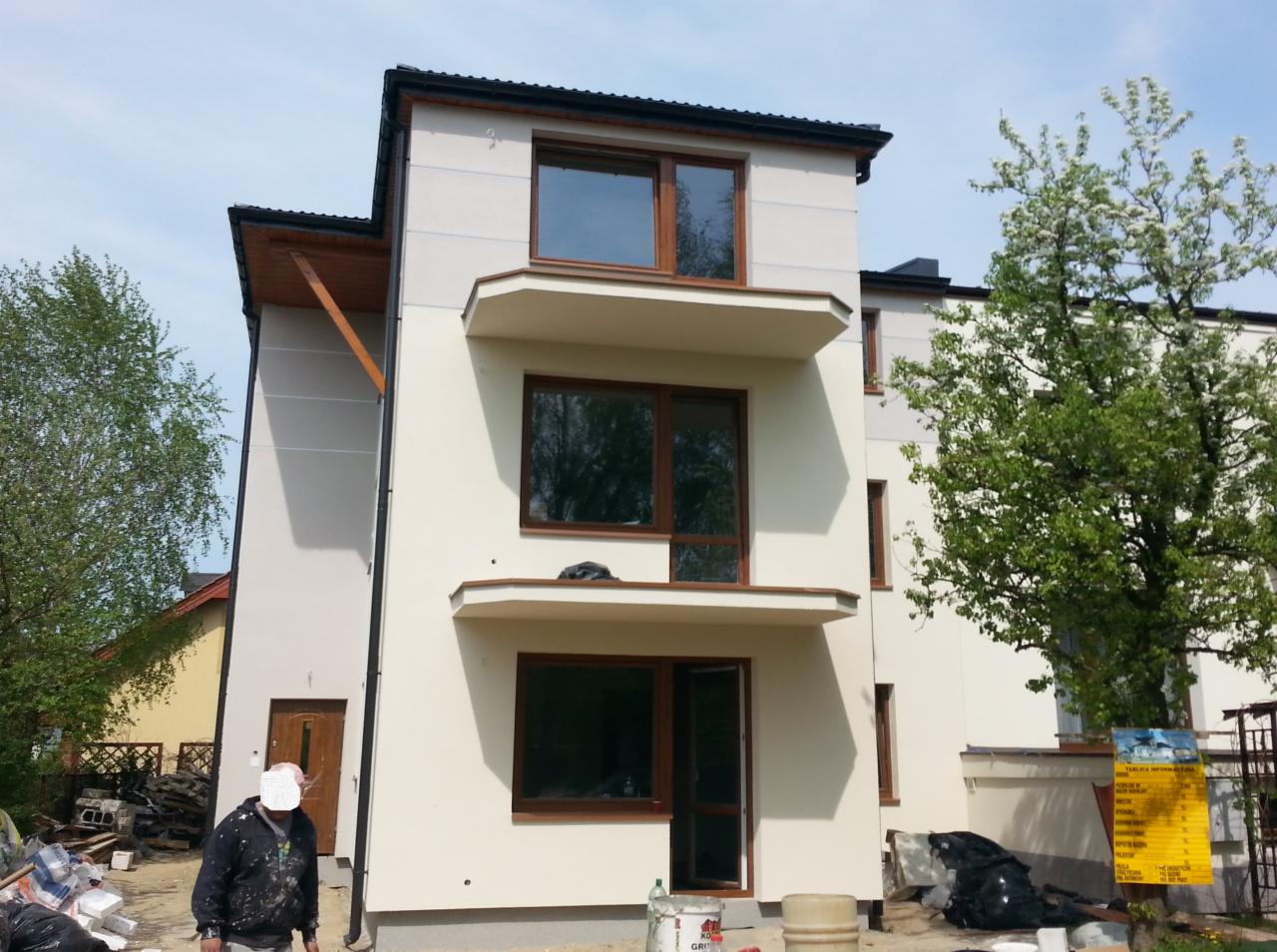 Dwupiętrowy budynek z jasną elewacją, ciemnymi ramami okiennymi i małymi balkonami, widoczny pracownik w ubraniu roboczym i materiały budowlane na pierwszym planie.