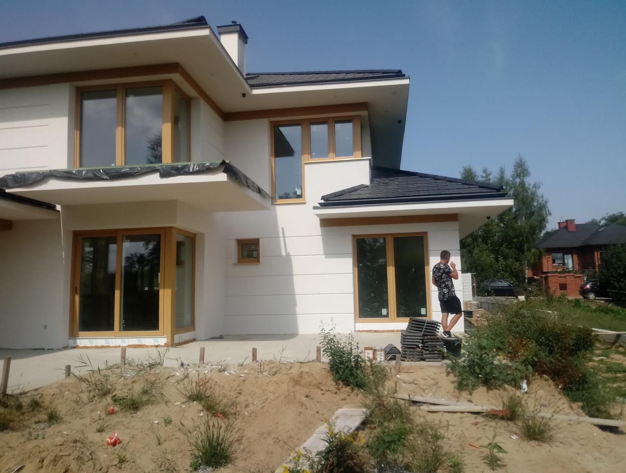 Nowoczesny dom jednorodzinny z jasną elewacją w trakcie wykończenia, widoczne okna z drewnianą stolarką, mężczyzna na zewnątrz w trakcie pracy, przygotowane płytki elewacyjne na ziemi.