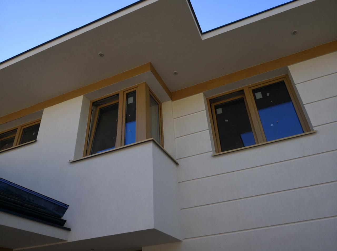 Nowoczesny dom z jasną elewacją, drewnianymi ramami okiennymi i minimalistycznym balkonem, widziany z dołu pod kątem, ukazujący detale wykończenia.