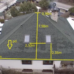 Widok z drona na dach domu pokryty grafitową dachówką bitumiczną z oknami dachowymi, oznaczony żółtymi liniami z wymiarami.