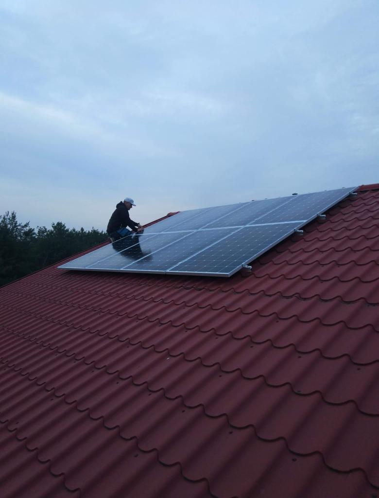 Instalacja paneli słonecznych na dachu pokrytym czerwoną dachówką w Boksyckiej, widok z dołu na montera w czarnej kurtce i białej czapce pracującego na wysokości.