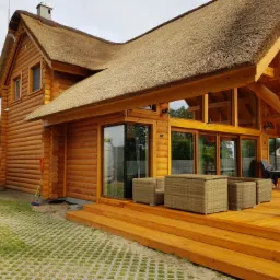 Drewniany dom z bali z trzcinowym dachem i tarasem, widoczny częściowo samochód dostawczy z otwartymi drzwiami i materiałami budowlanymi w środku.