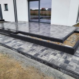 BRUKI GORZELAK - Nowoczesny taras z kostki brukowej w odcieniach szarości z czarnymi obrzeżami, z widocznymi donicami na rośliny i fragmentem białego budynku w tle. Powierzchnia kostki lekko wilgotna, odbijająca...