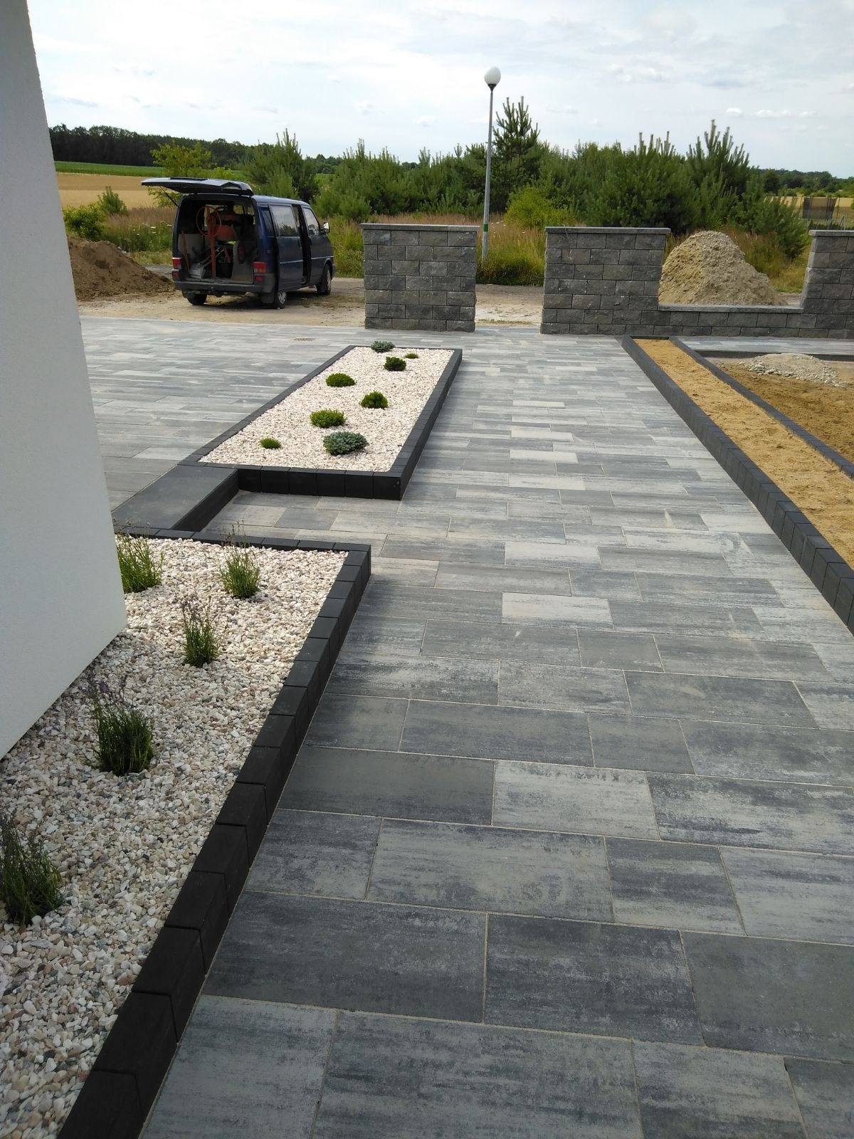 Ułożona szara kostka brukowa z czarnym obrzeżem, rabaty z białym żwirem i niskimi krzewami ozdobnymi, częściowo ukończony murek z kostki, samochód dostawczy w tle.