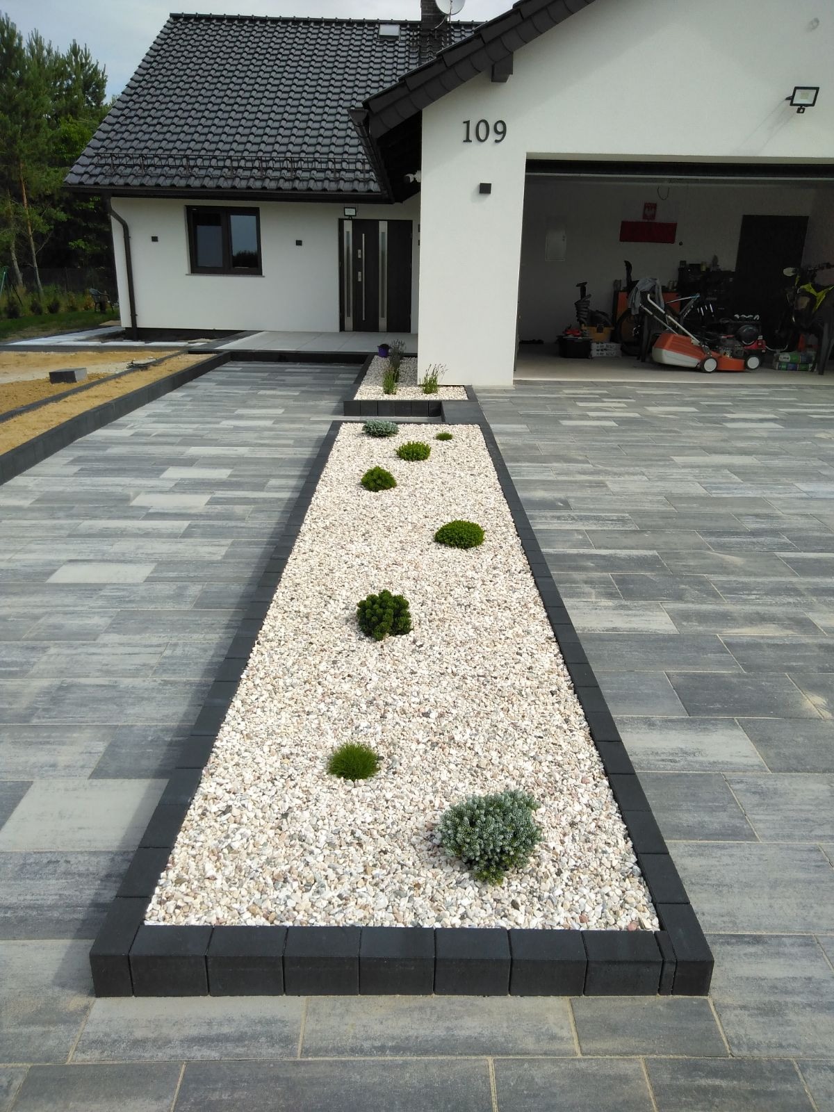 Nowoczesny podjazd do domu z szarej kostki brukowej, obrzeża z czarnej kostki, pas z białego grysu i nasadzeniami niskich roślin ozdobnych.