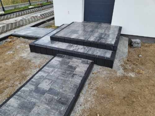 Świeżo ułożona ścieżka i schody z szarej kostki brukowej prowadzące do drzwi wejściowych budynku, widoczne niedociągnięcia w postaci piasku na kostce i niezakończone prace wykończeniowe.