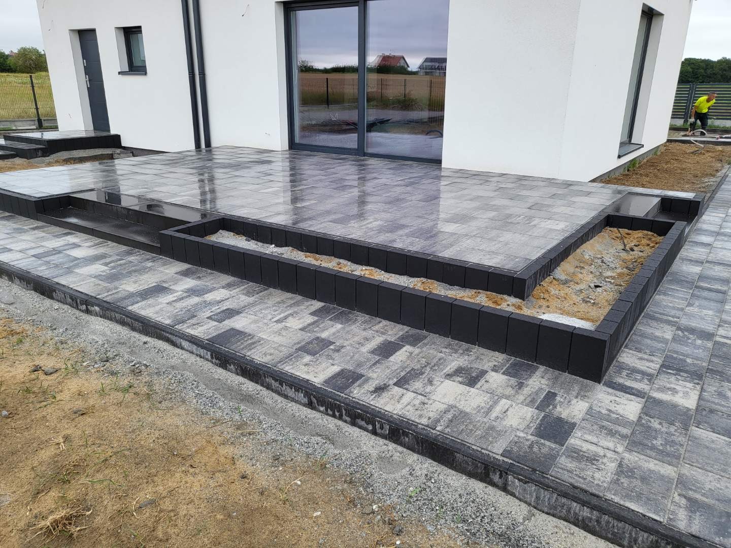 Nowoczesny taras z kostki brukowej w odcieniach szarości z czarnymi obrzeżami, z widocznymi donicami na rośliny i fragmentem białego budynku w tle. Powierzchnia kostki lekko wilgotna, odbijająca...