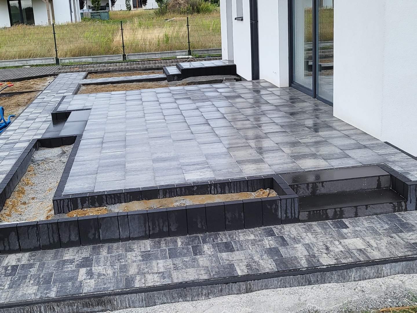 Nowoczesny taras z szarych płyt betonowych w trakcie budowy, z elementami rabat kwiatowych obłożonych czarną kostką, widok z góry na częściowo ukończony projekt.