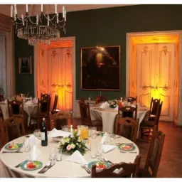 Elegancka sala balowa z okrągłymi stołami nakrytymi białymi obrusami, kwiatami, zastawą i oświetleniem w ciepłych barwach, podkreślającym dekoracyjne drzwi i sztukaterie.