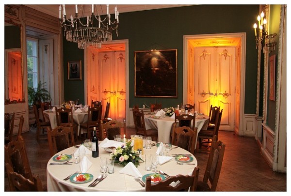 Elegancka sala balowa z okrągłymi stołami nakrytymi białymi obrusami, kwiatami, zastawą i oświetleniem w ciepłych barwach, podkreślającym dekoracyjne drzwi i sztukaterie.