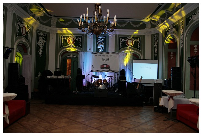 Sala balowa z parkietem przygotowana na występ zespołu muzycznego: scena z perkusją, instrumentami, nagłośnieniem, oświetleniem punktowym, projektorem i banerem 'We are the champions'.