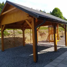 Drewniana konstrukcja z dachem w Węgierskiej Górce, prawdopodobnie zadaszenie tarasu lub wiata, z poziomymi listwami tworzącymi ściany, ustawiona na żwirowym podłożu, z widocznym systemem rynnowym.