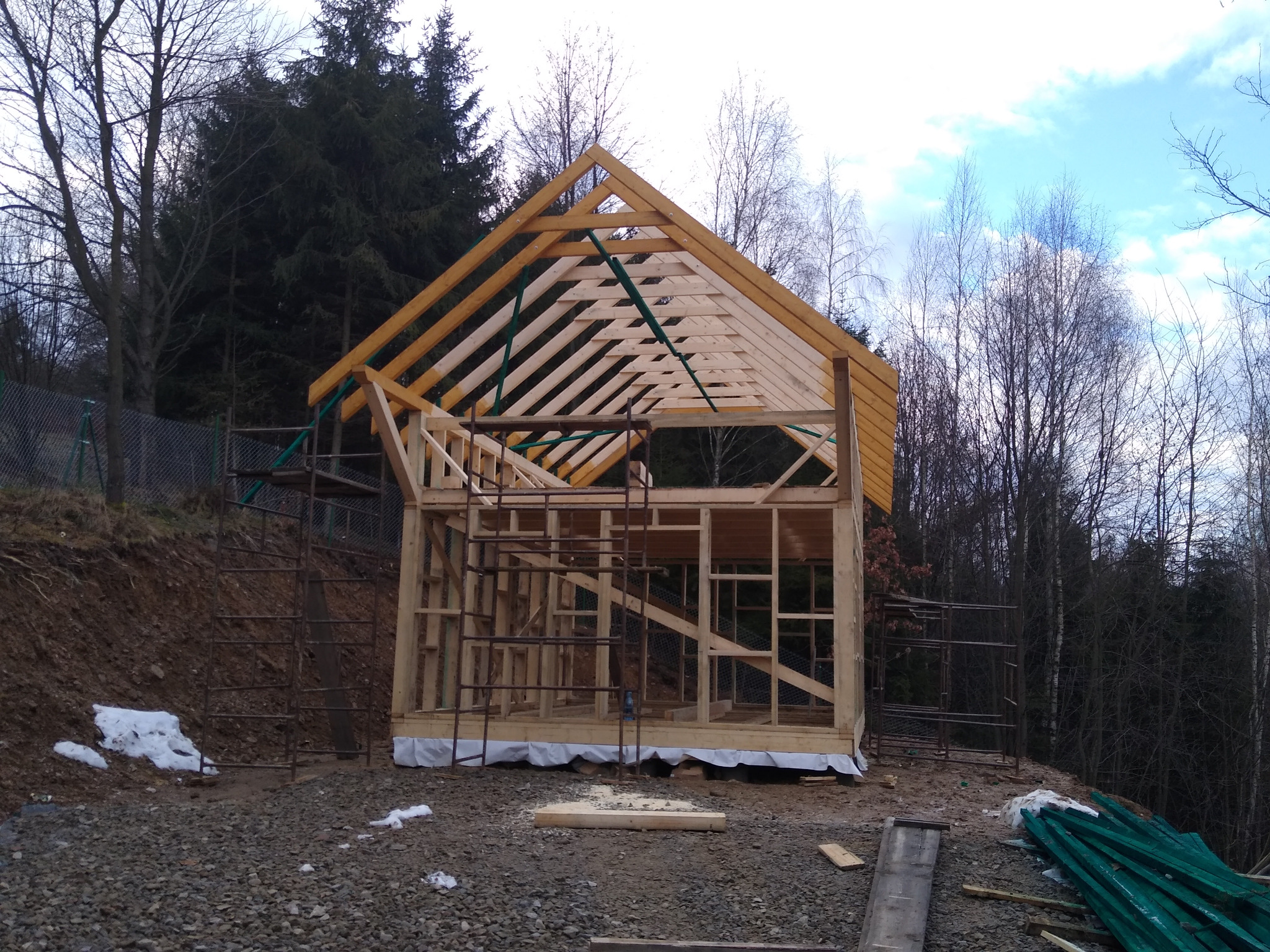 Surowa konstrukcja szkieletowa domu z widoczną więźbą dachową na tle drzew i pochmurnego nieba. Widoczne rusztowania i elementy konstrukcyjne.