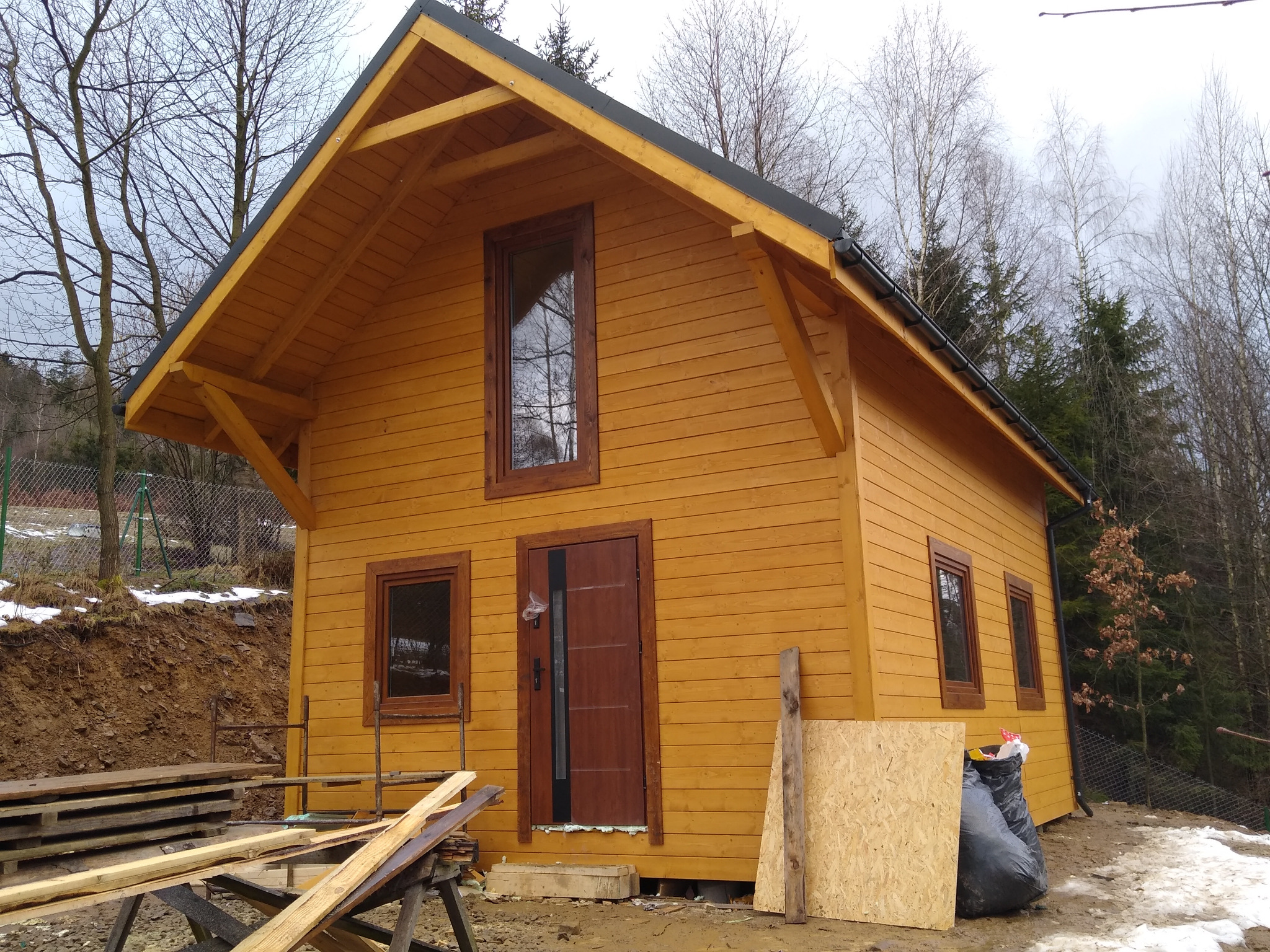 Żółty, drewniany dom szkieletowy z brązowymi oknami i drzwiami, widoczny z zewnątrz, z elementami budowlanymi w otoczeniu. Częściowo ośnieżony teren.