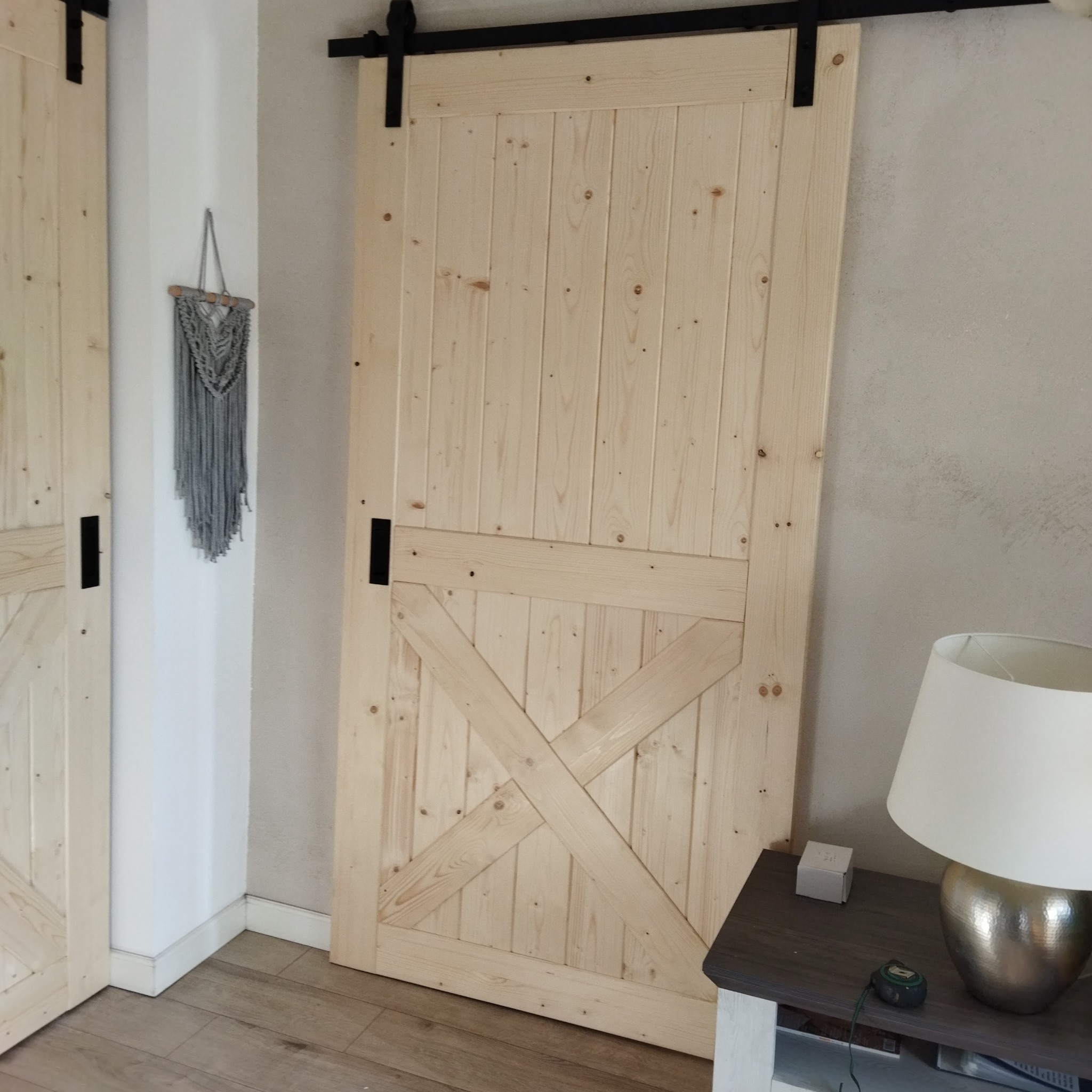 Jasne, drewniane drzwi przesuwne w stylu rustykalnym z czarnymi okuciami, zamontowane na szynie. W tle widoczny fragment drugiej pary drzwi i dekoracja ścienna.