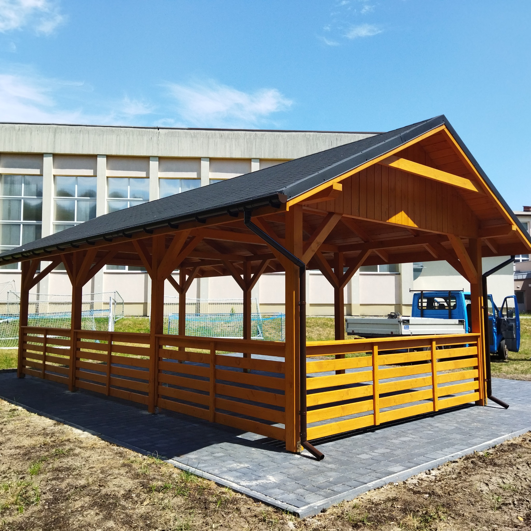 Drewniana altana z czarnym dachem na kostce brukowej, z widocznym fragmentem budynku w tle. Uwagę zwraca intensywny kolor drewna i staranne wykonanie detali.