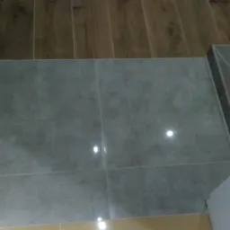 Szare płytki podłogowe ułożone w przejściu z drewnianej podłogi, widoczne fragmenty białej ściany i dolnej części ściany w kolorze beżowym.