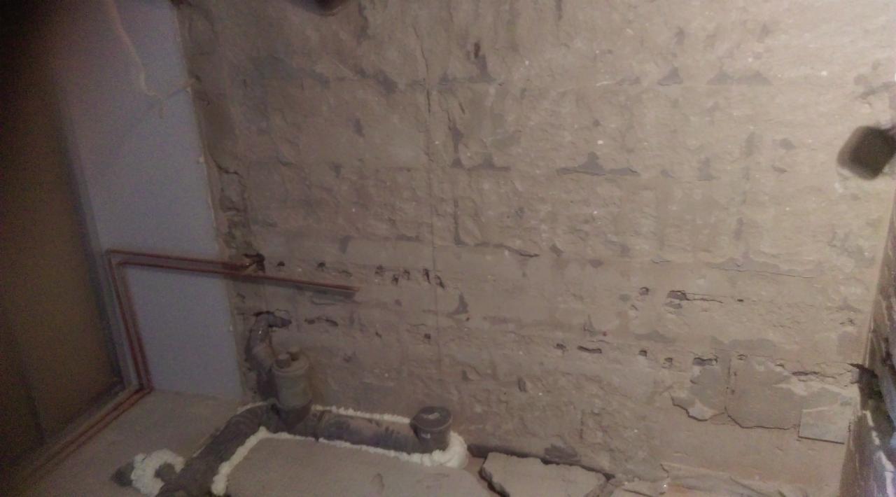Surowa ściana z widocznymi śladami po skuciu płytek, fragment instalacji hydraulicznej przy podłodze i pionowa rura w rogu pomieszczenia.
