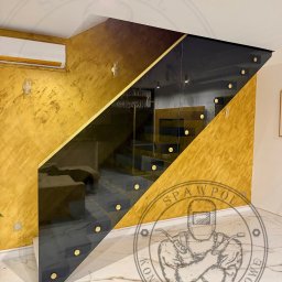 Spawpol Arkadiusz Polerowicz - Nowoczesne schody z balustradą szklaną, złote akcenty na stopniach i ścianie, marmurowa podłoga. Widok na salon przez szklaną balustradę.