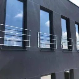 Zewnętrzne, metalowe zabezpieczenia okienne, wykonane z prostych, poziomych prętów, zamontowane na elewacji budynku o ciemnoszarym, surowym wykończeniu.