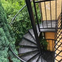 Zewnętrzne schody spiralne z czarnego metalu prowadzące na mały balkon, otoczone bujną zielenią.