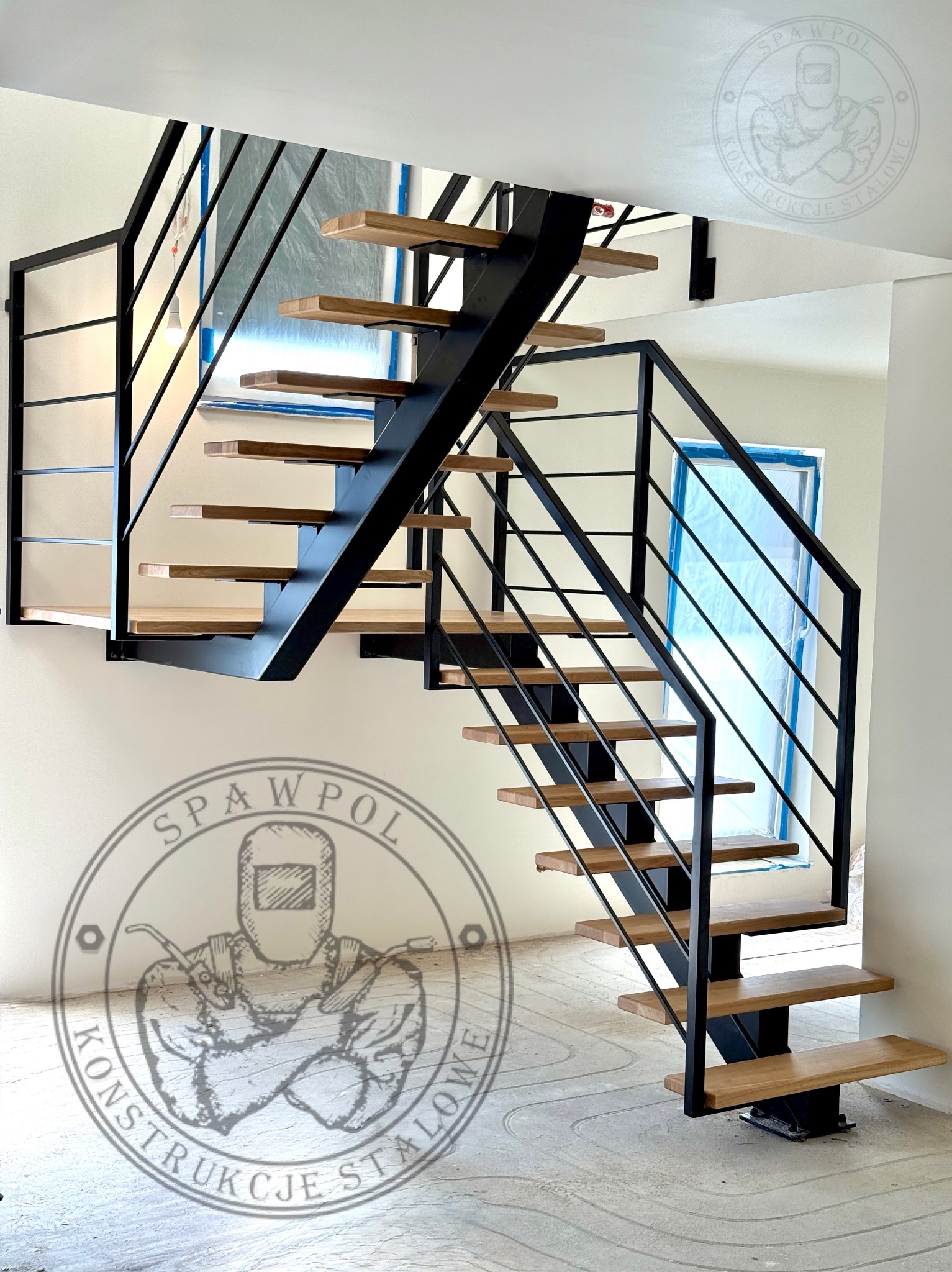 Nowoczesne schody metalowe z drewnianymi stopniami, czarna konstrukcja, minimalistyczny design, widok z dołu, logo firmy na ścianie.