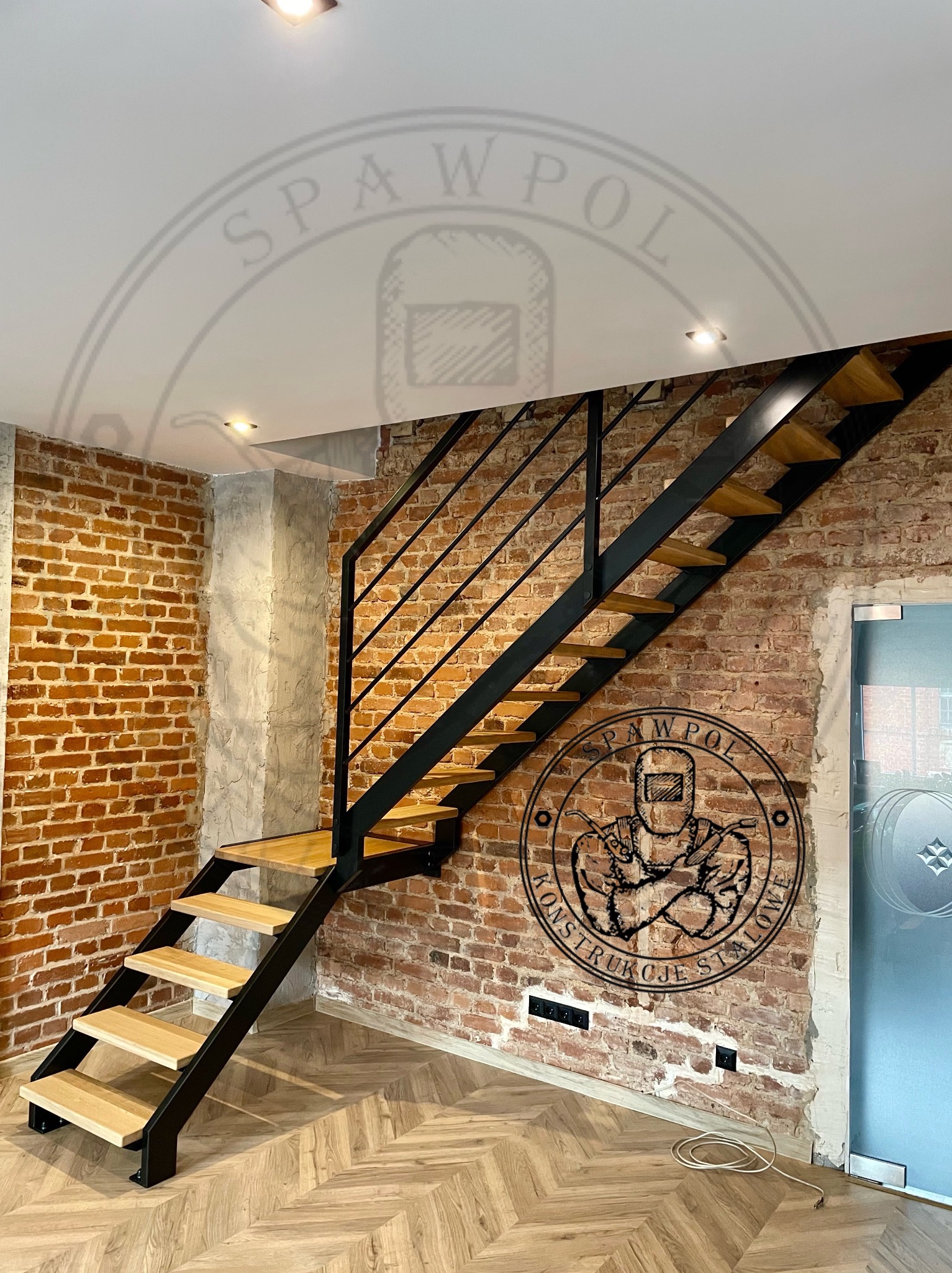 Nowoczesne schody metalowe z drewnianymi stopniami, czarna konstrukcja, ceglana ściana w tle, parkiet w jodełkę. Widoczne logo firmy na ścianie.