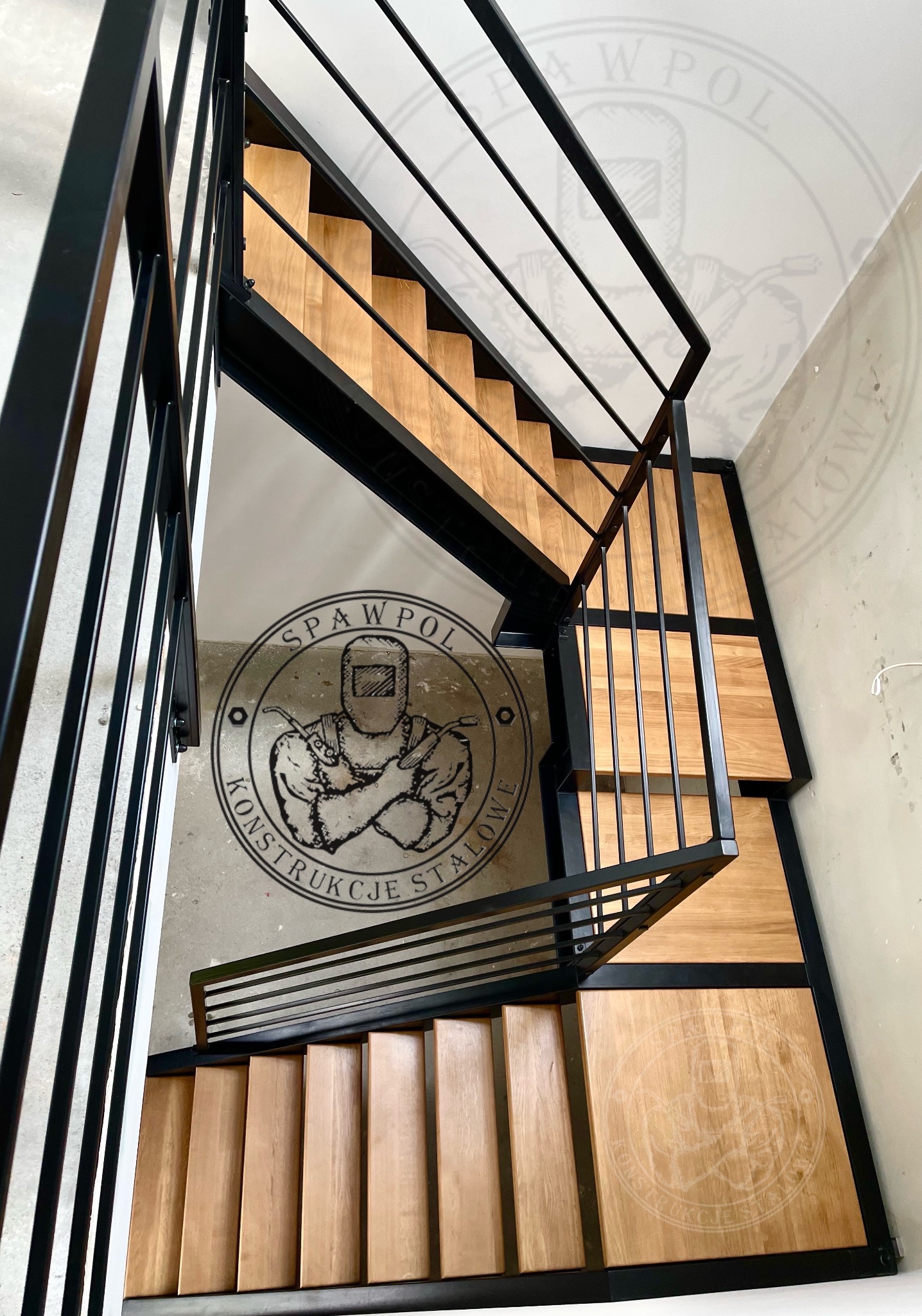 Nowoczesne schody z drewnianymi stopniami i czarną metalową konstrukcją, widok z góry, z logo firmy na ścianie. Minimalistyczny design.
